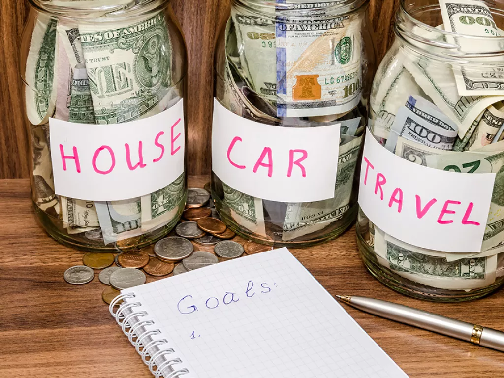 jars labeled house, car, and travel with money in them and a notebook that says goals