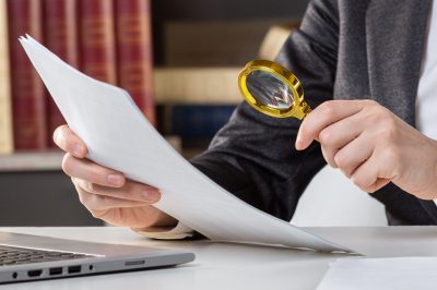 Female hands with a golden loupe and a papers at the desktop next to open laptop, close up, selected focus.