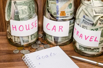 jars labeled house, car, and travel with money in them and a notebook that says goals