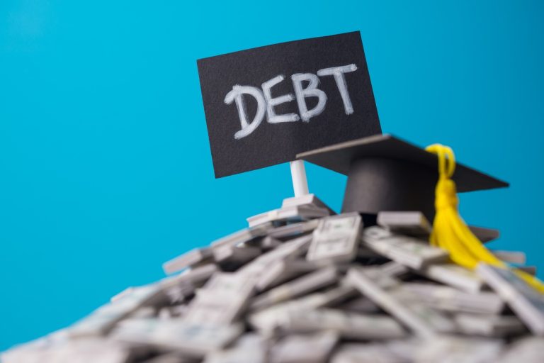 Mortarboard on heap US dollar banknotes and DEBT label with blue background. Student debt or loan, education, studying, investment for future, student financial aid and repayment planning concept.