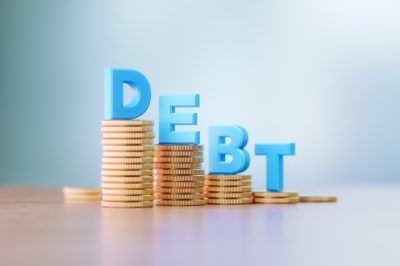 Blue capital letters sitting on coins stacks write debt before defocused background.