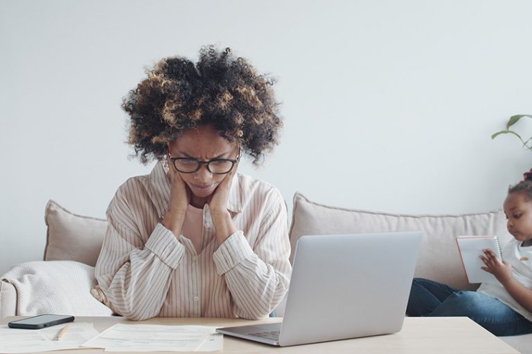 a woman sits at a table with a laptop in front of her, head in her hands, while her child plays behind her