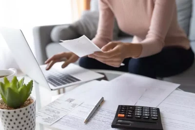 image of a desk scattered with papers, a laptop, and a calculator as a woman types something into the laptop